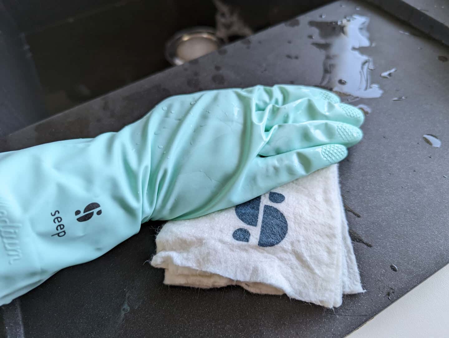 A hand wearing natural rubber gloves by seep uses an all purpose cloth by Seep to wipe down a black granite sink