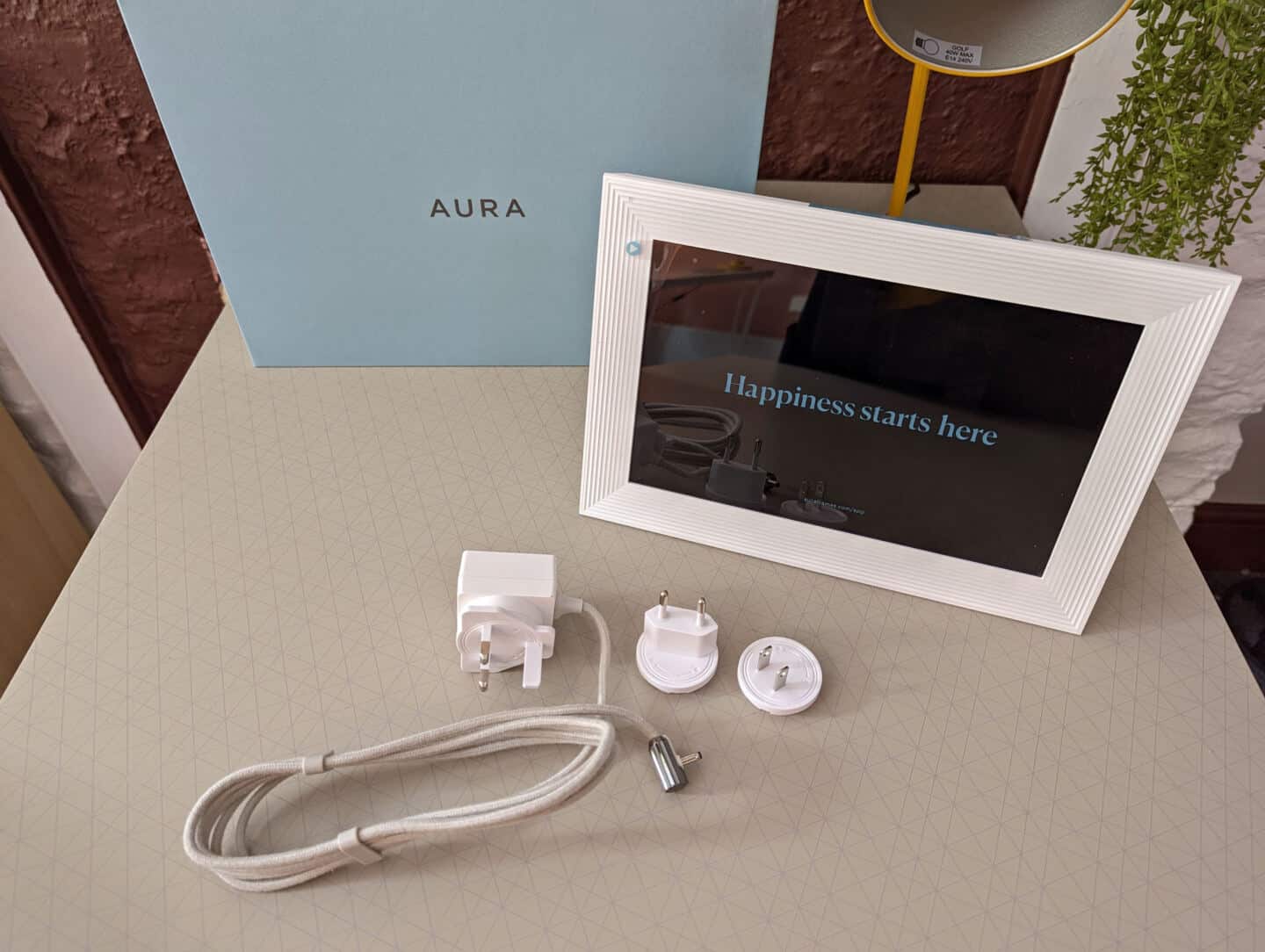 A blue box and a digital photo frame with power cable is on a desk next to a yellow lamp.