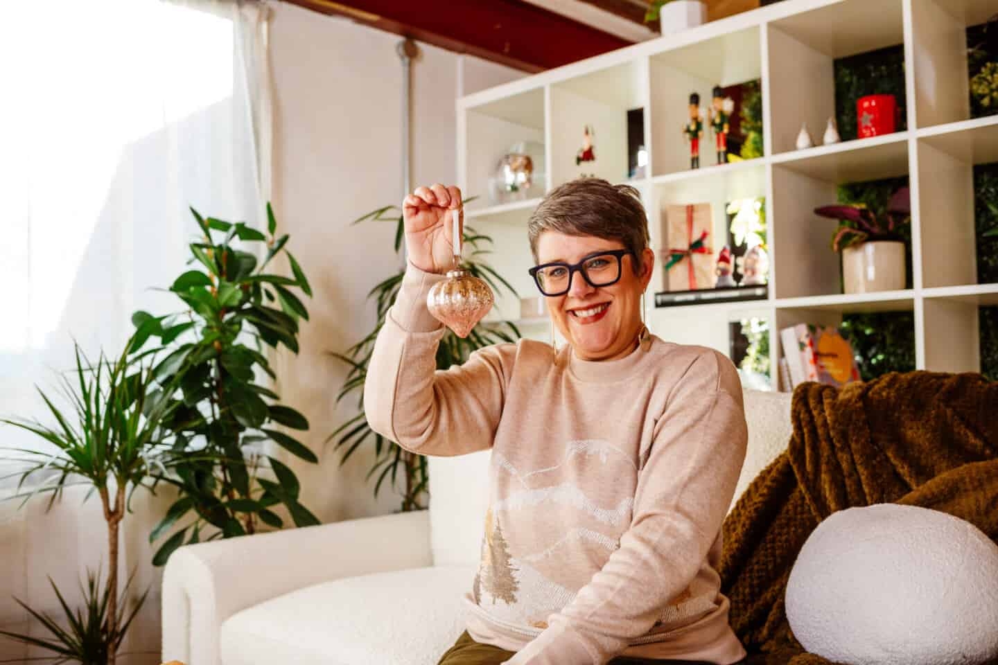 A woman in her forties sat on a sofa holding up a Christmas bauble from Balsam Hill