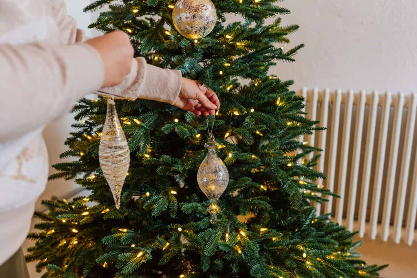 A woman in her forties hangs Christmas baubles on a realistic artificial Christmas tree from Balsam Hill