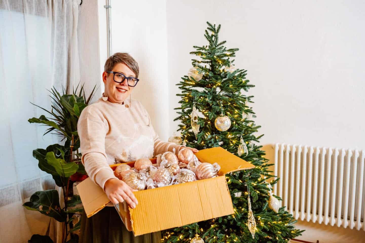 A woman in her forties holds a box of Christmas baubles in front of a realistic artificial Christmas tree from Balsam Hill