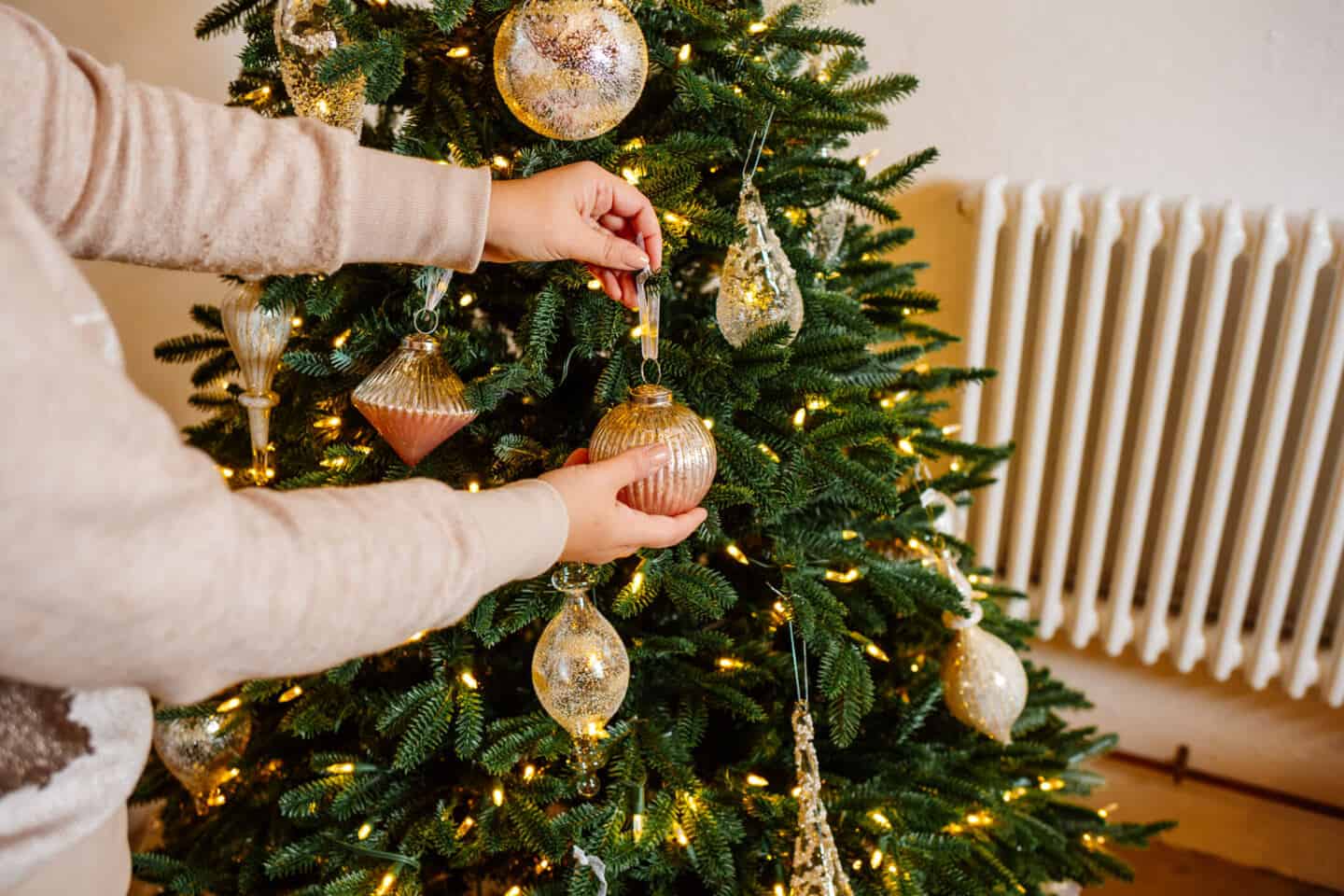 A woman in her forties hangs Christmas baubles on a realistic artificial Christmas tree from Balsam Hill