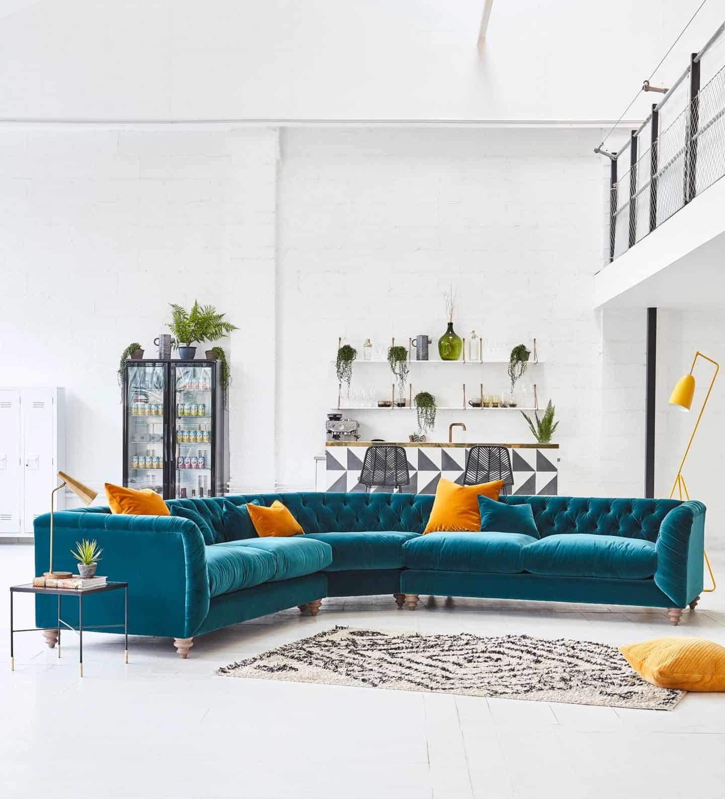 A large turquoise velvet corner sofa from Darlings of Chelsea with mustard yellow scatter
cushions sits in a warehouse setting. Furniture can be found on furniture comparison website ufurnish.com