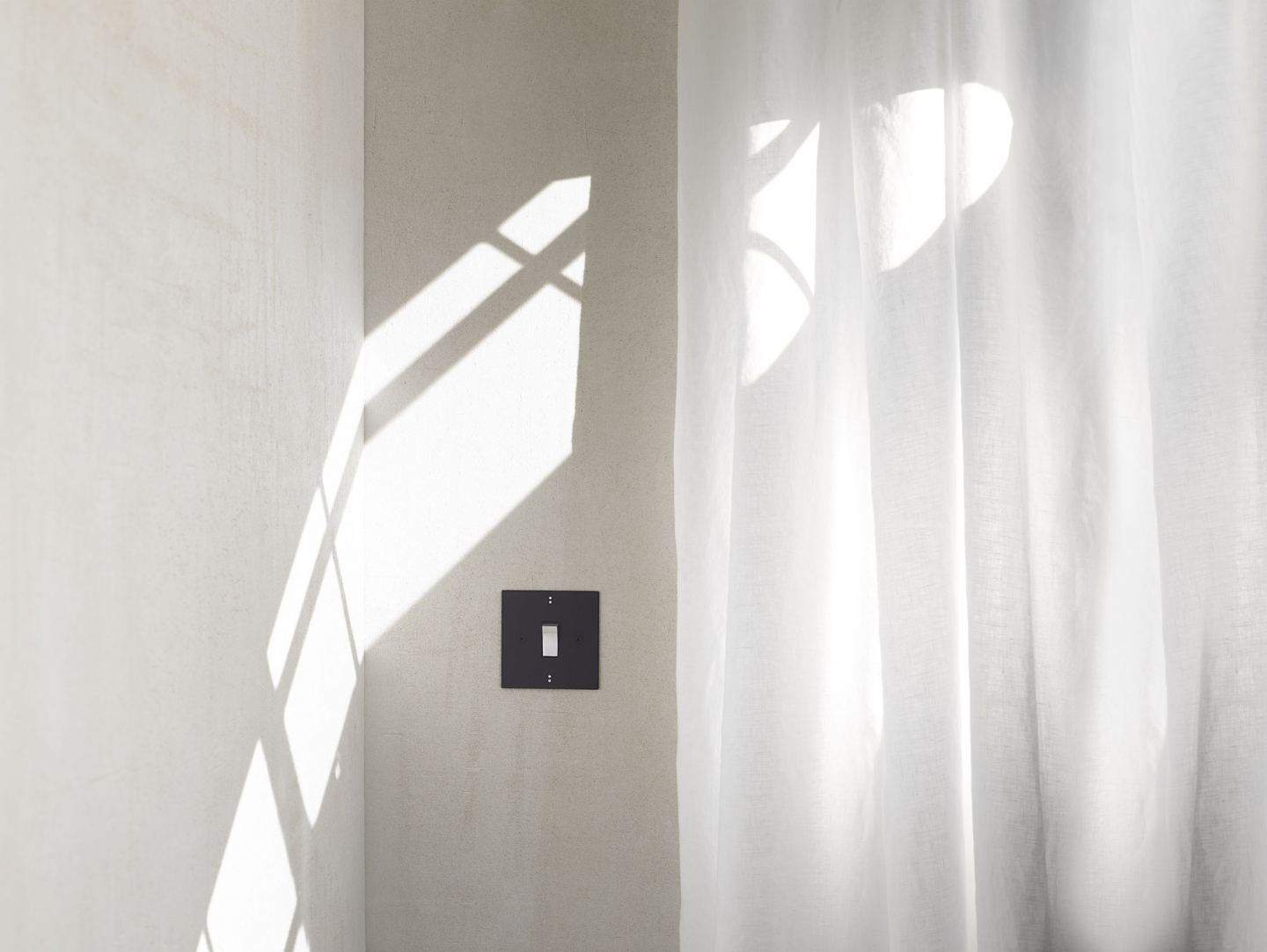 Matt black designer light switch on the wall beside a floaty linen curtain