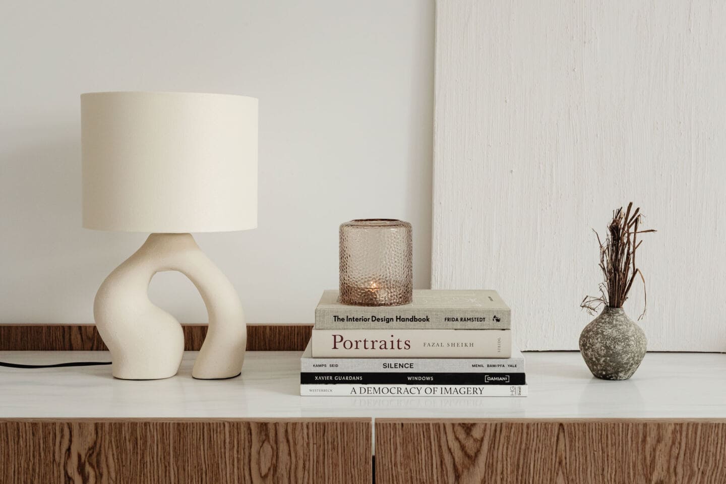 An organic shaped lamp in a sideboard next to a pile of books and some dried twigs in a small vase