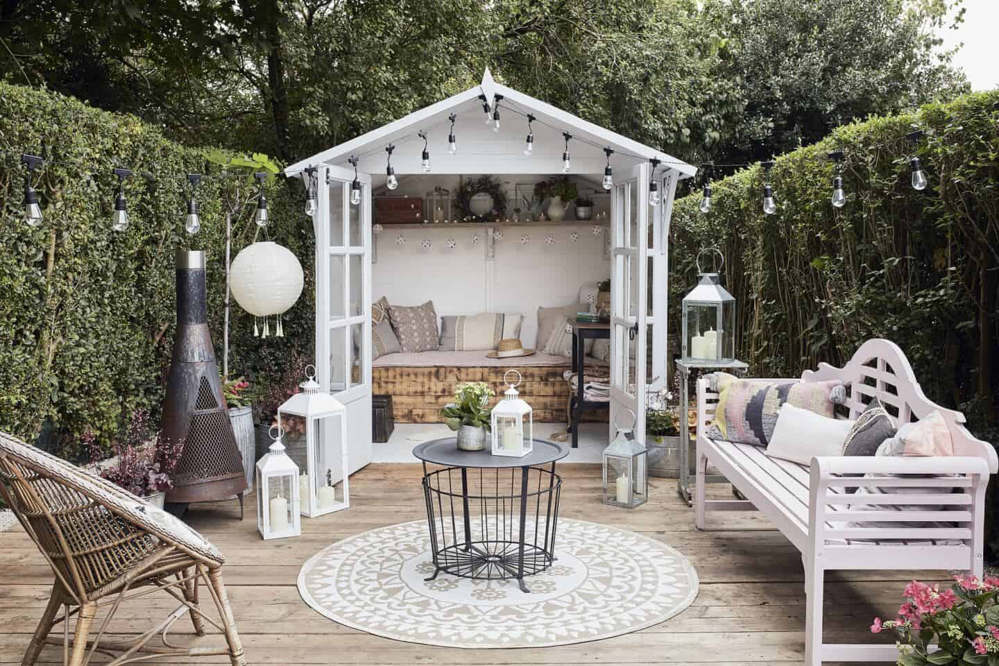 A garden summer house at the bottom of the garden. A small seating area leads off from it with chairs, benches, a chimnea, festoon lights and some hurricane lanterns.
