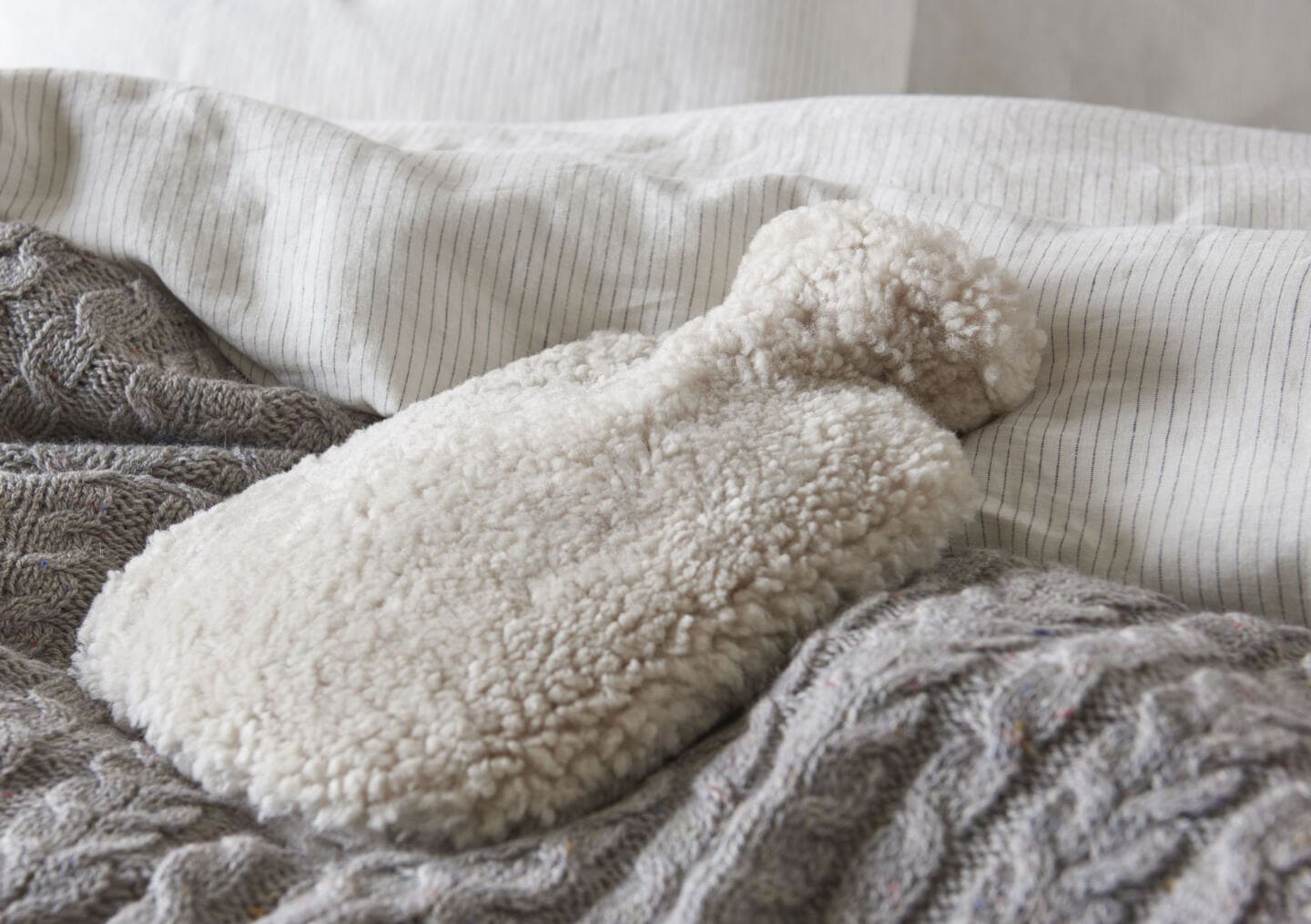 A hot water bottle in a a fluffy white cover laid on a grey woollen blanket on a bed