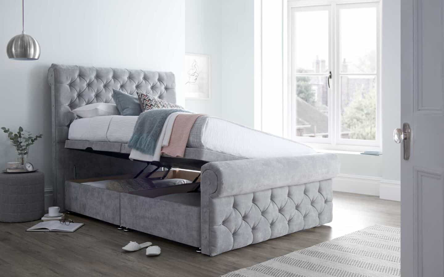 An open grey upholstered ottoman bed in a bright an airy bedroom with large windows