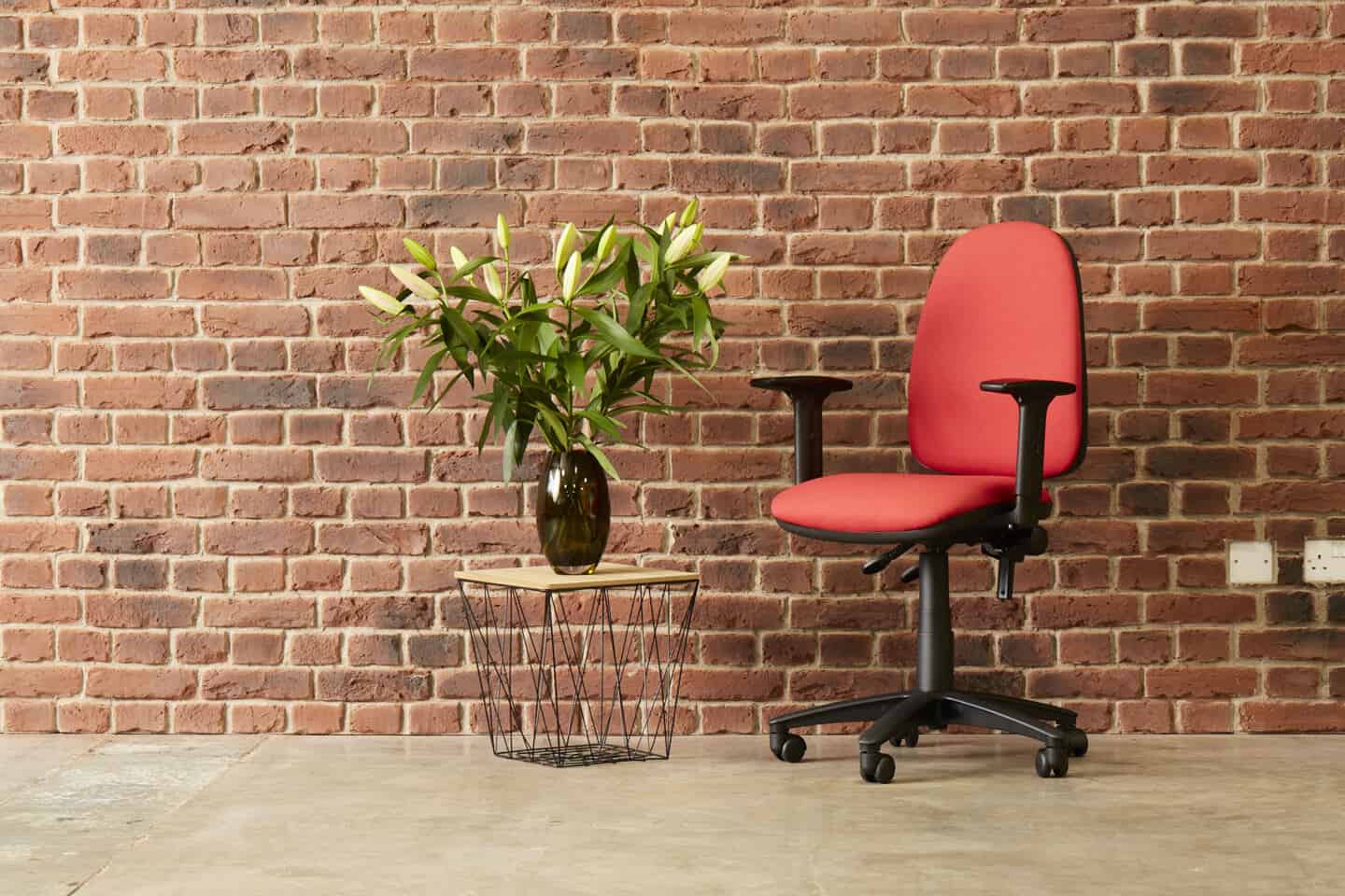 An ergonomic office chair infront of an exposed brick wall and next to a coffee table with a vase of flowers on it