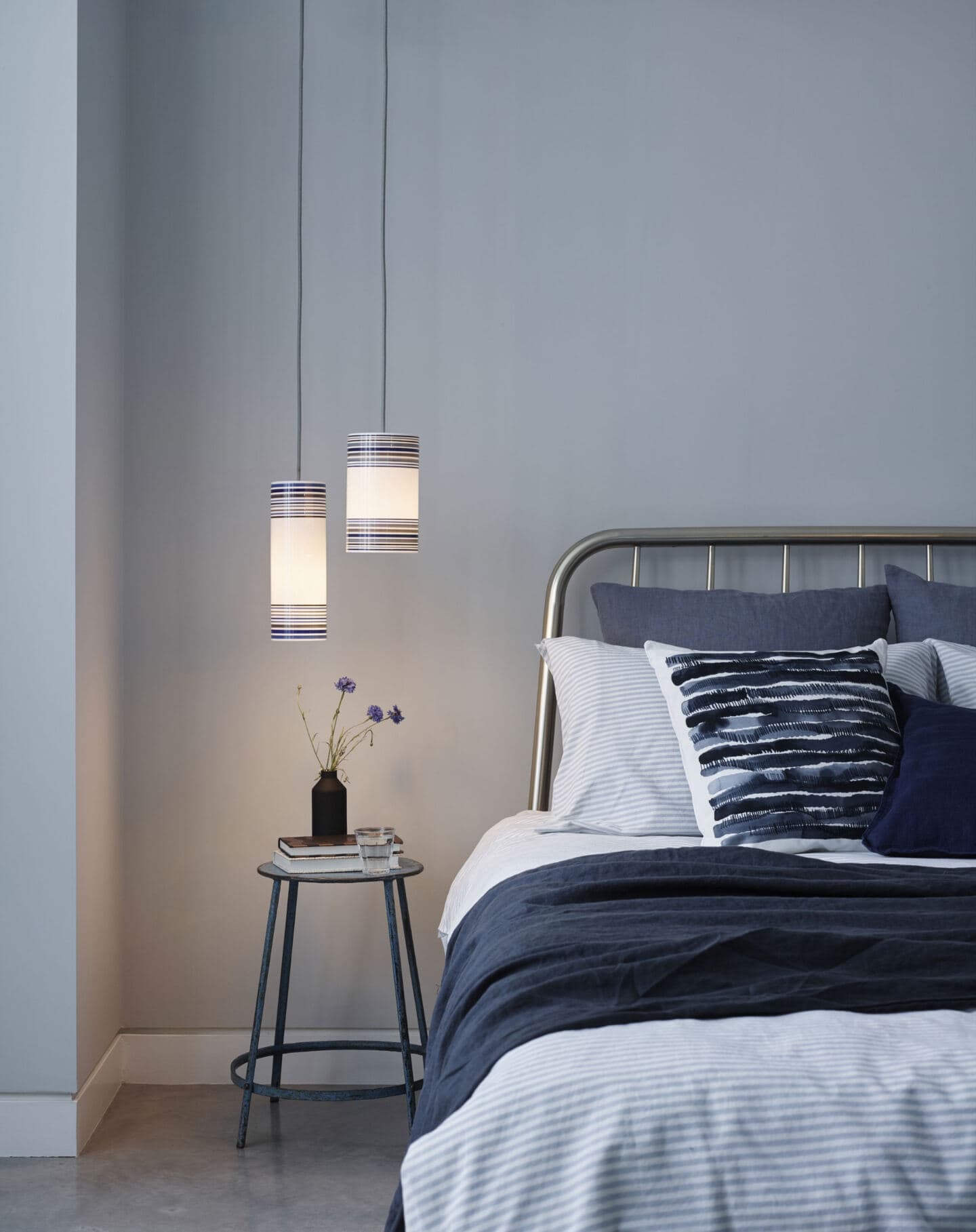 Blue bedroom featuring a metal bed and side table and bedding in natural materials. Tow lights hang down from ceiling above side table