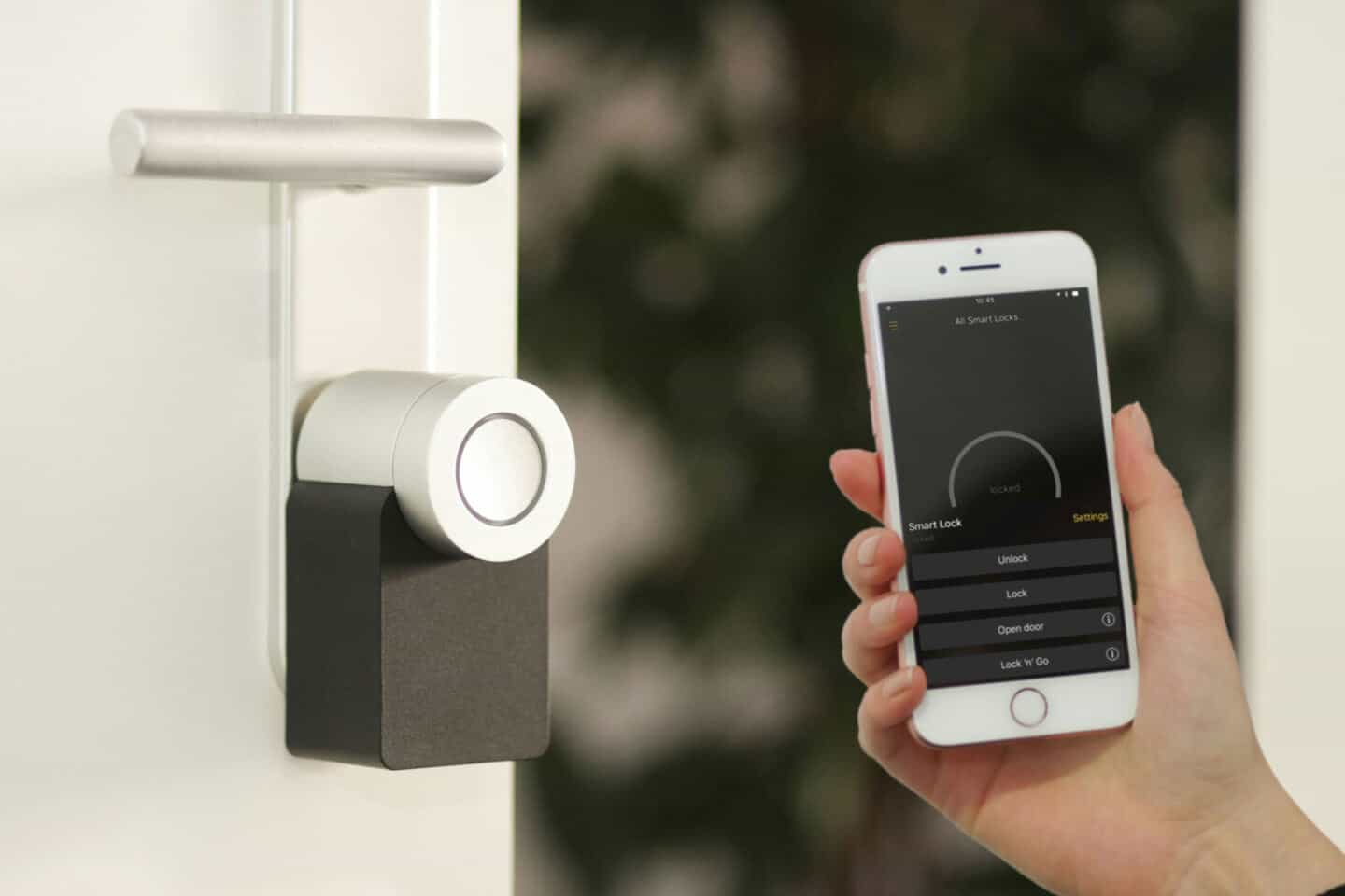 A woman holds a mobile phone with a home security app on it next to a smart lock on a door
