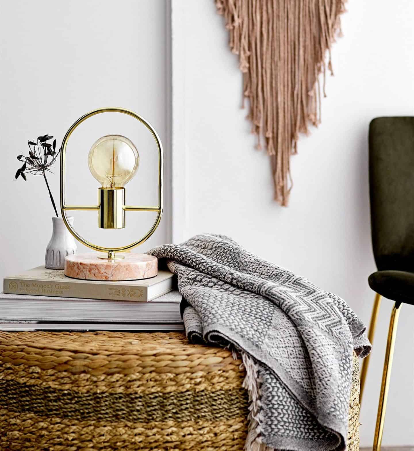Shekåbba The Danish Home stocks a range of Danish homewares. A table lamp sat on a stack of books next to a grey throw on a rattan pouffe.
