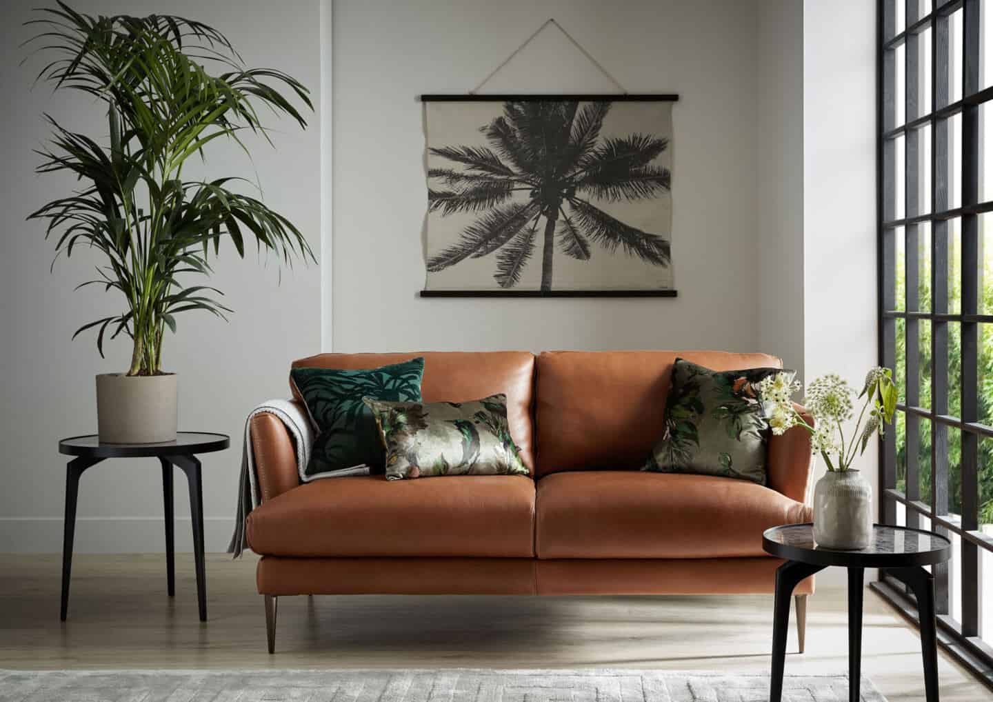 Bringing Natural Elements into the Home. Living room and featuring a brown leather sofa with botanical cushions. A botanical wall hanging is on the wall behind and views through the window.