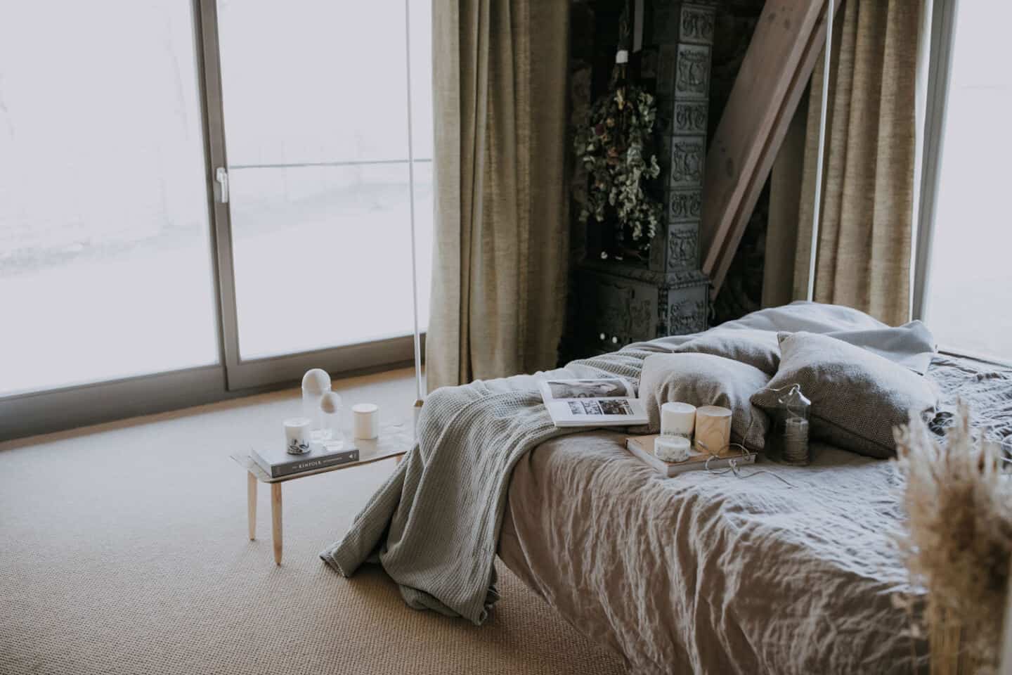 Textured Lives. Using texture in interiors to bring your home to life. A bedroom with floor to ceiling wondows and natural light pouring in. The bed has a linen cover and there are books, candles and cushions on the bed.