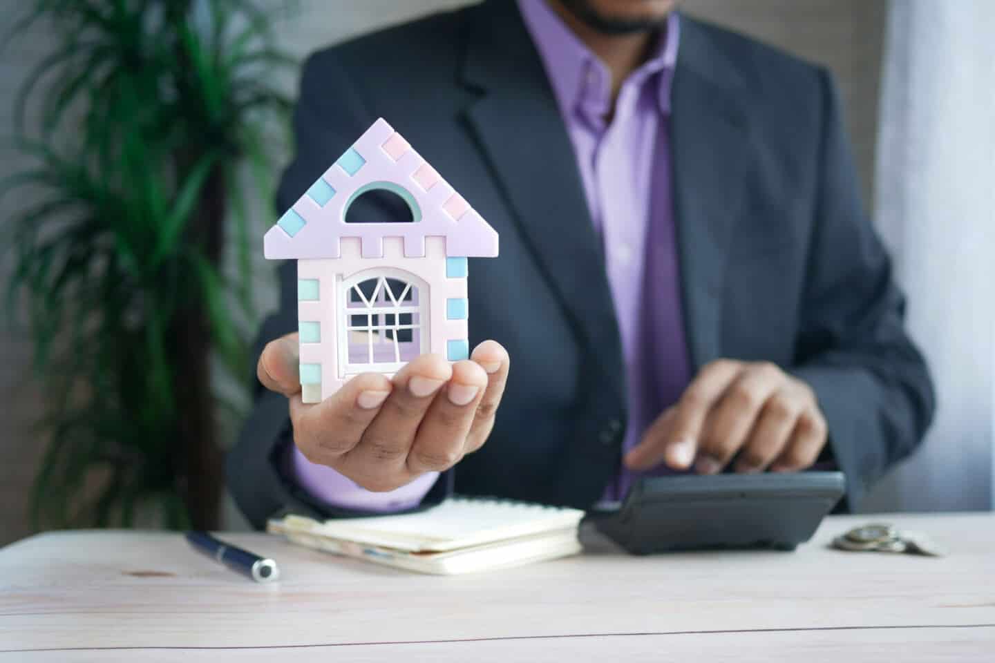 A man holding a small plastic home in one hand and using a calculator in the other.