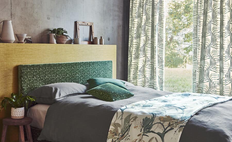 Made-to-Measure Curtains & Blinds. A bed in front of floor to ceiling window. Long geometric curtains in Villanova fabric hang behind the bed which is covered in a bed runner.