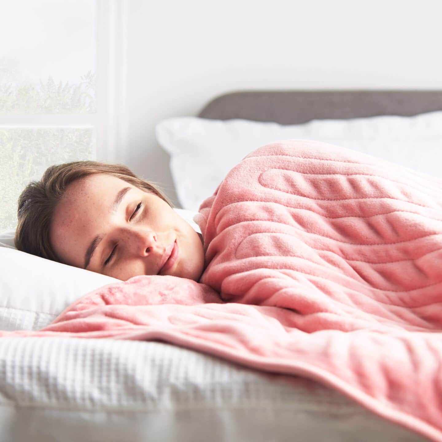 A white woman in her thirties asleep under a pink electric blanket
